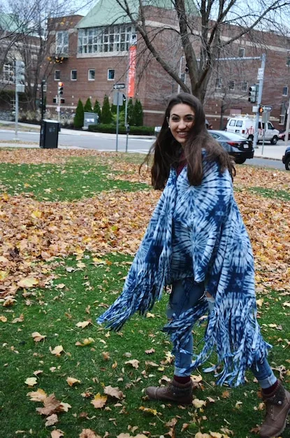 A woman standing outdoors wearing a large, patterned scarf draped over her shoulders as a shawl, paired with a simple black dress and ankle boots.