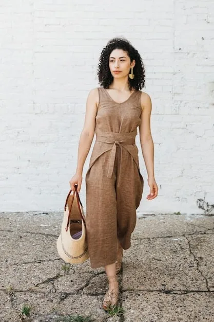 A young woman wearing a beige midi linen dress with a fitted bodice and flared skirt, accessorized with a wide-brimmed straw hat and brown leather sandals in a cobblestone street.