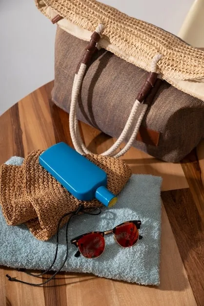 A close-up of a straw tote bag with leather handles, filled with a linen scarf, sunglasses, and a water bottle, sitting on a sandy beach.