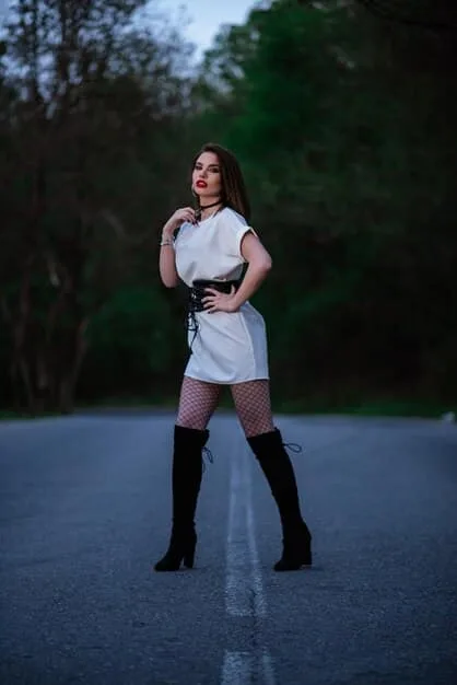 A petite woman standing in a well-lit studio, wearing black heeled over-the-knee boots, a black mini skirt, and a fitted white turtleneck sweater. She is posing confidently with her hand on her hip.