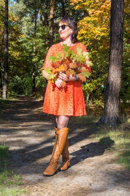 A woman standing in a park during autumn, wearing beige flat over-the-knee boots, a long-sleeved black sweater dress, and a brown fedora hat. Colorful fall foliage surrounds her.