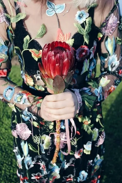A close-up shot of a runway look featuring a model wearing a bold floral print dress. The dress has large, vibrant flowers in shades of pink, orange, and green. The background is blurred, emphasizing the intricate details of the floral design.