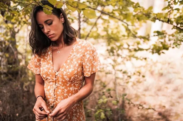 A woman with warm skin tones wearing an olive green dress, accessorized with gold jewelry and a tan leather belt. The background is a sunlit outdoor setting.