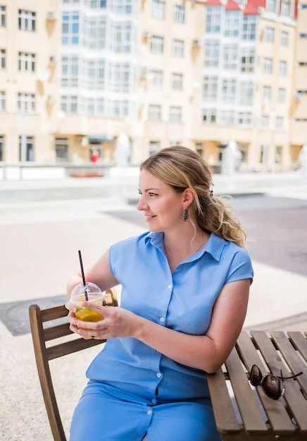 A person is sitting at an outdoor cafe, wearing a light blue button-down shirt with the sleeves rolled up, paired with khaki chinos and brown leather loafers. They are sipping coffee and reading a book.