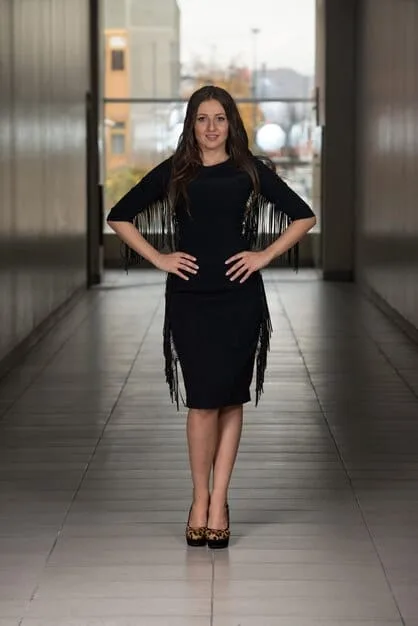 A woman is shown from the waist up, wearing a simple black dress. In one image, she's wearing a thin, understated leather belt for daytime. In the other, she's wearing a wider belt with a decorative gold buckle for evening. The background is a plain, neutral color.