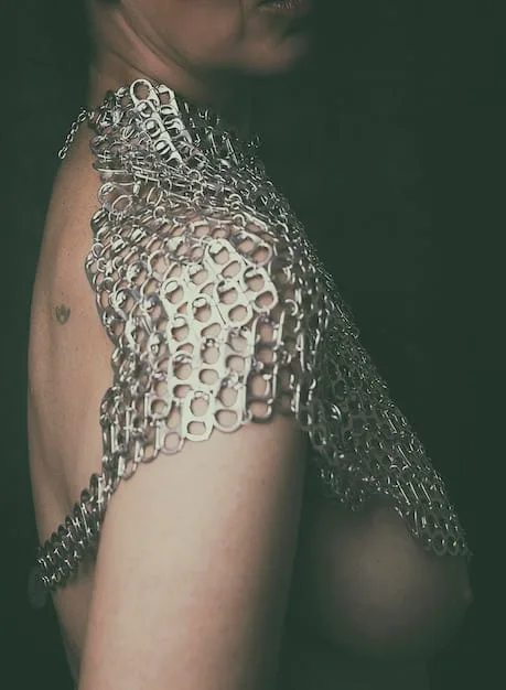 A close-up of a woman's torso showcasing a unique statement belt made of interwoven metallic chains and leather. The belt cinches a flowing, neutral-toned dress at the waist, accentuating the silhouette. The background is minimalistic to draw attention to the belt's intricate details and texture.