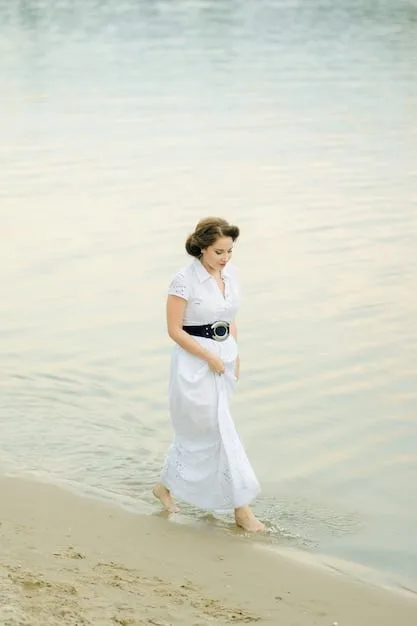 A woman in a flowing, white linen maxi dress, accessorized with a wide-brimmed straw hat and delicate gold jewelry, walking barefoot on a sandy beach during a daytime wedding ceremony.