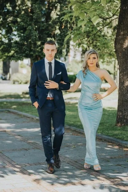 A couple dressed in cocktail attire at a garden wedding; the woman is wearing a navy blue lace dress with nude heels, and the man is wearing a light grey suit with a patterned tie.