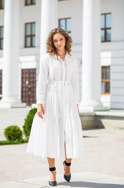 A person stylishly dressed in a capsule wardrobe outfit consisting of a white flowing midi skirt and a black tee; the model is wearing black wayfarer sunglasses, black leather sandals and stands in a trendy neighborhood with minimalistic aesthetics.