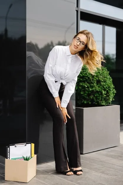 A woman wearing a classic white button-down shirt with sleeves rolled up, paired with tailored black trousers and simple gold jewelry. She is standing in a bright, modern office space.