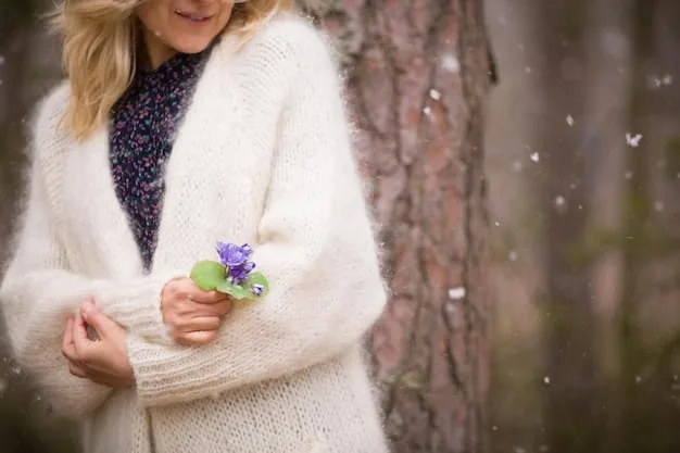 A woman standing in a park, wearing a cream-colored chunky knit sweater and a floral midi skirt. She is accessorized with ankle boots and a crossbody bag, creating a stylish and comfortable outfit. The background features lush greenery and soft natural light, emphasizing the relaxed yet chic vibe.