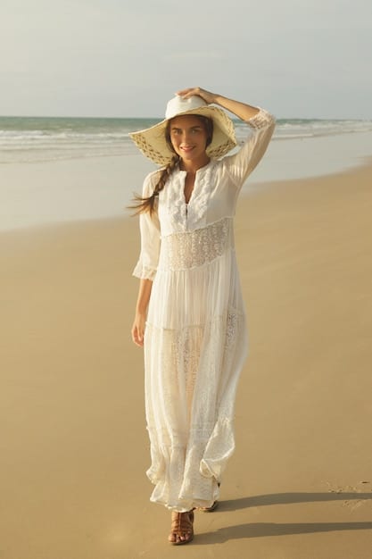A woman standing on a beach during sunset, wearing a flowy white dress layered with a light, embroidered kimono and accessorized with a wide-brimmed hat and sunglasses.