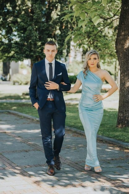 A couple dressed in cocktail attire at a garden wedding; the woman is wearing a navy blue lace dress with nude heels, and the man is wearing a light grey suit with a patterned tie.