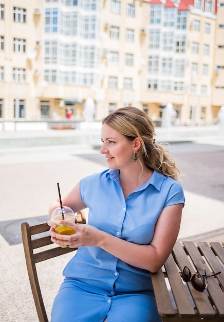 A person is sitting at an outdoor cafe, wearing a light blue button-down shirt with the sleeves rolled up, paired with khaki chinos and brown leather loafers. They are sipping coffee and reading a book.