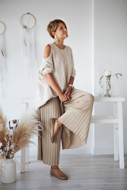 A person with neutral skin tone wearing a muted blush-colored dress, paired with a light gray cardigan and nude heels. The background is a soft, diffused studio lighting.