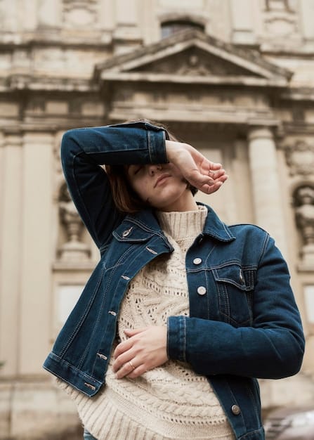 A street style shot showcases a woman wearing a dark wash denim jacket paired with light wash denim jeans. She is wearing a white t-shirt underneath and accessorizing with brown leather boots and a matching belt, creating a contrasting yet cohesive double denim look. The background is a bustling city street with a mix of modern and historic buildings.
