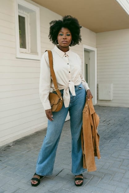 A model is wearing wide-leg, high-waisted dark wash jeans with a tucked-in cream-colored silk blouse. She is accessorizing with pointed-toe heeled boots and a structured handbag, showcasing a sophisticated and polished way to style wide-leg denim. The background is a clean, minimalist studio setting.
