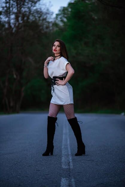 A petite woman standing in a well-lit studio, wearing black heeled over-the-knee boots, a black mini skirt, and a fitted white turtleneck sweater. She is posing confidently with her hand on her hip.