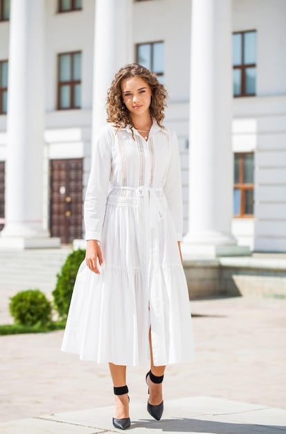 A person stylishly dressed in a capsule wardrobe outfit consisting of a white flowing midi skirt and a black tee; the model is wearing black wayfarer sunglasses, black leather sandals and stands in a trendy neighborhood with minimalistic aesthetics.