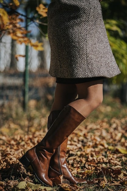 A close-up shot of a woman's legs wearing knee-high suede boots in a rich caramel color. She is standing on a cobblestone street with fallen leaves around her feet. The focus is on the texture and detail of the boots, highlighting the craftsmanship and quality.