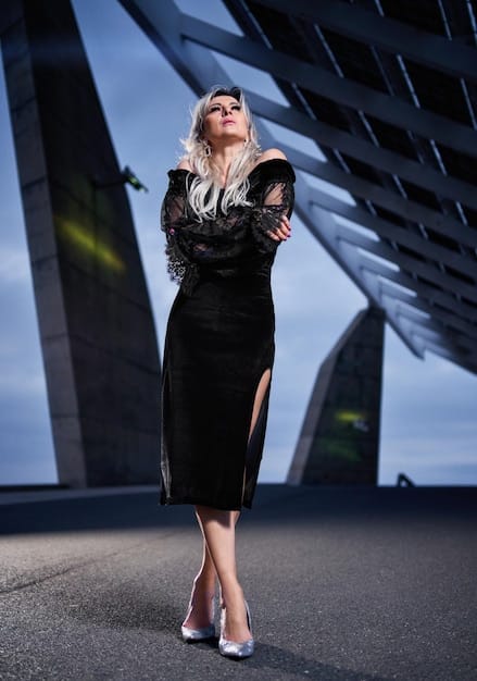 A woman in a sleek, black midi dress with a high slit, paired with silver strappy heels and a bold red lip, standing against a minimalist, modern backdrop.