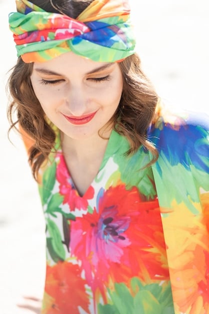 A woman wearing a flowing, painterly floral print dress in a sunlit studio. The dress has a mix of vibrant and muted colors, with brushstroke-like patterns. The overall look is artistic and bohemian.
