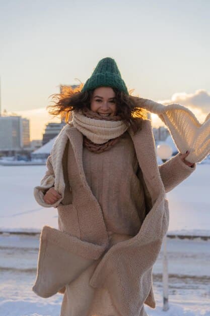 A person wearing a stylish oversized wool coat in a neutral color (e.g., camel, gray, or cream), paired with a cozy knit scarf and leather gloves, standing in a snowy urban setting.