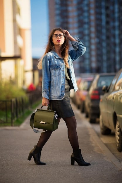 A woman wearing a navy blazer over a white t-shirt and jeans, paired with ankle boots. She is holding a leather handbag and smiling confidently. The background is an urban street scene.