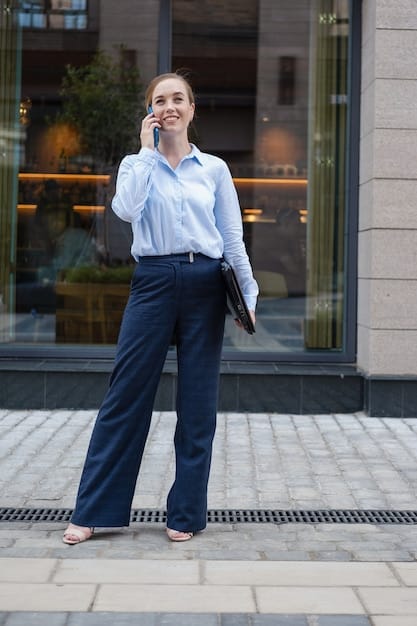 A woman wearing a business casual outfit: dark blue blazer, light blue button-down shirt, dark grey chinos, and brown loafers. She is standing in a modern office setting.