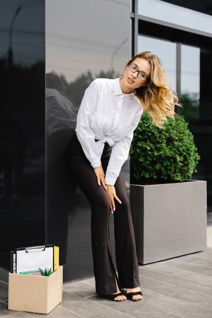 A woman wearing a classic white button-down shirt with sleeves rolled up, paired with tailored black trousers and simple gold jewelry. She is standing in a bright, modern office space.