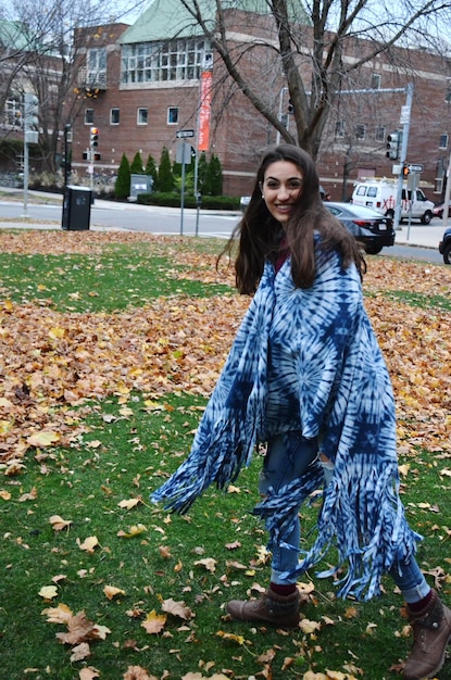 A woman standing outdoors wearing a large, patterned scarf draped over her shoulders as a shawl, paired with a simple black dress and ankle boots.